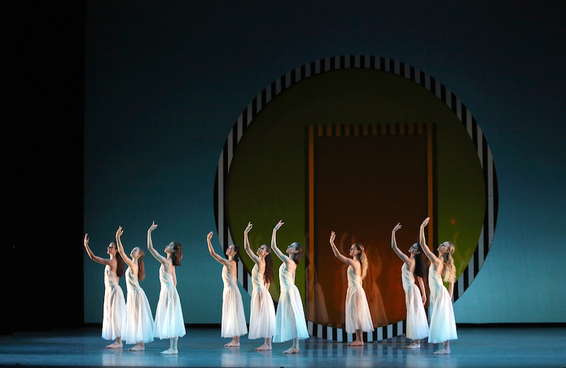 A group of women in Grecian white dresses stand with feet together. Their left arm curves overhead.